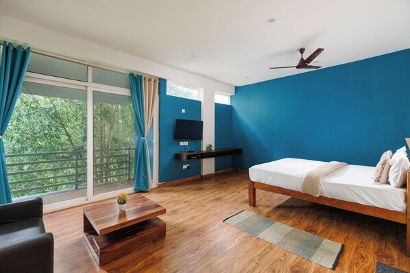 A bedroom with a bed, a TV, a wooden coffee table, and a view of greenery through the window. - Bougainvillea by Stayjade Poolbonfire Mysore Rd (Channapatna)