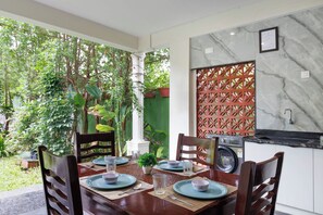 A dining area with a table set for four, surrounded by chairs, with a view of greenery outside. - Bougainvillea by Stayjade Poolbonfire Mysore Rd (Channapatna)