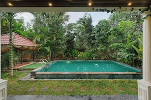A rectangular swimming pool with a tiled edge, surrounded by lush greenery and a covered seating area with a red roof. - Bougainvillea by Stayjade Poolbonfire Mysore Rd (Channapatna)