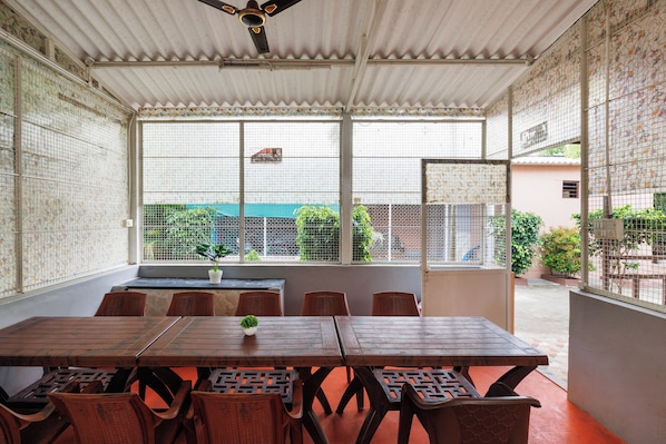 A covered outdoor dining area with a table and chairs, large windows with a view of the street, and a ceiling fan.