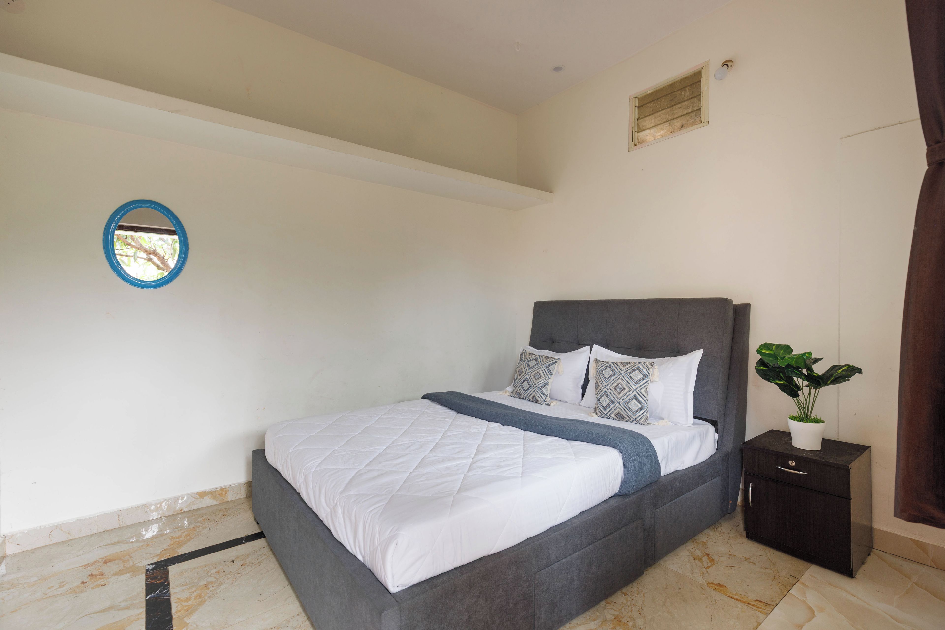 Un dormitorio con cama, mesita de noche, planta en maceta y espejo montado en la pared.