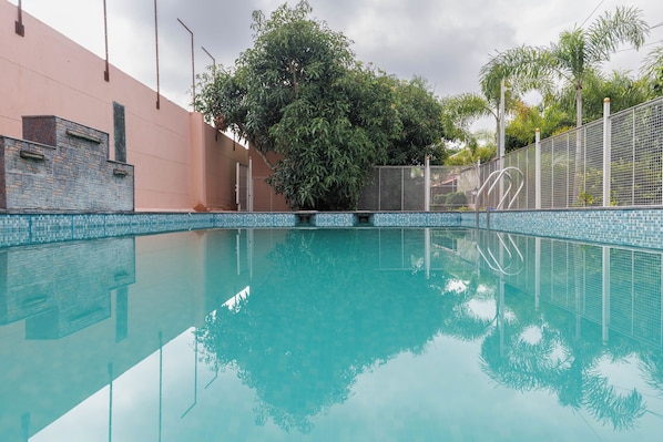 A clear swimming pool with a tiled edge, surrounded by a fence and trees. - White Swan by Stayjadepoolbbqbonfire Mysore Rd (Ramanagara)