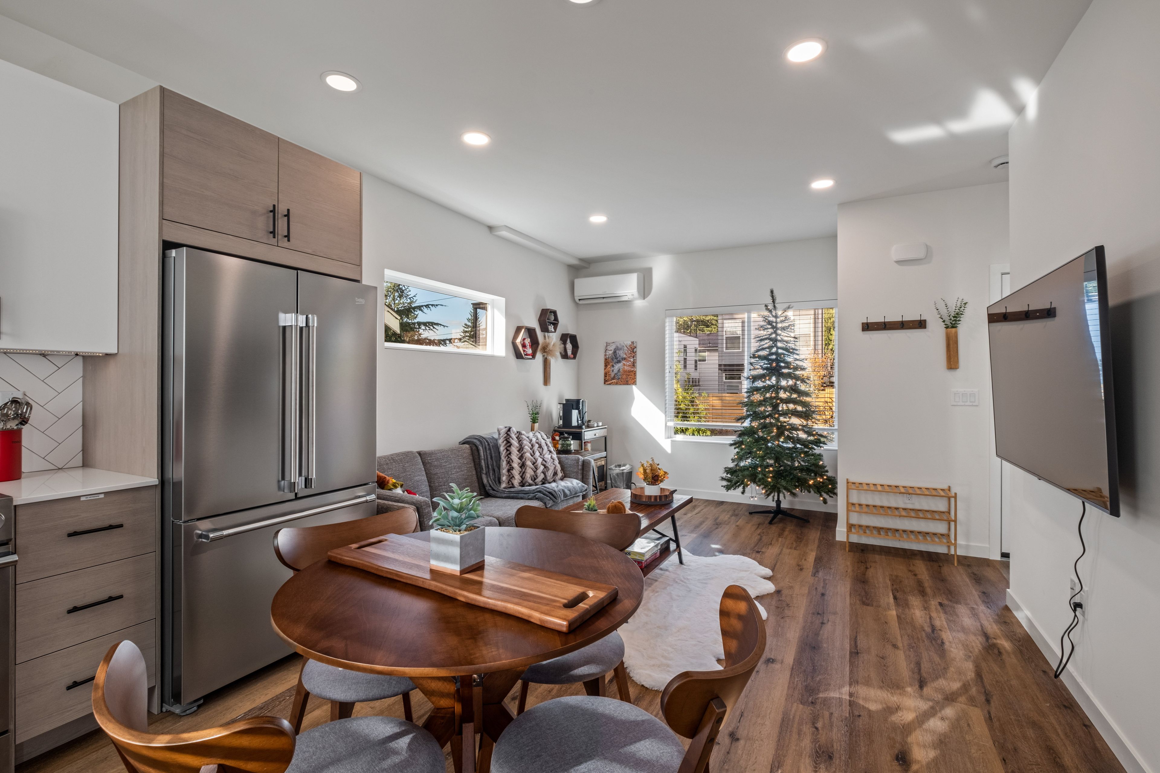 Une cuisine moderne dotée d’un réfrigérateur encastré, un coin repas avec une table et des chaises en bois, et un espace de vie comprenant un canapé et un sapin de Noël.