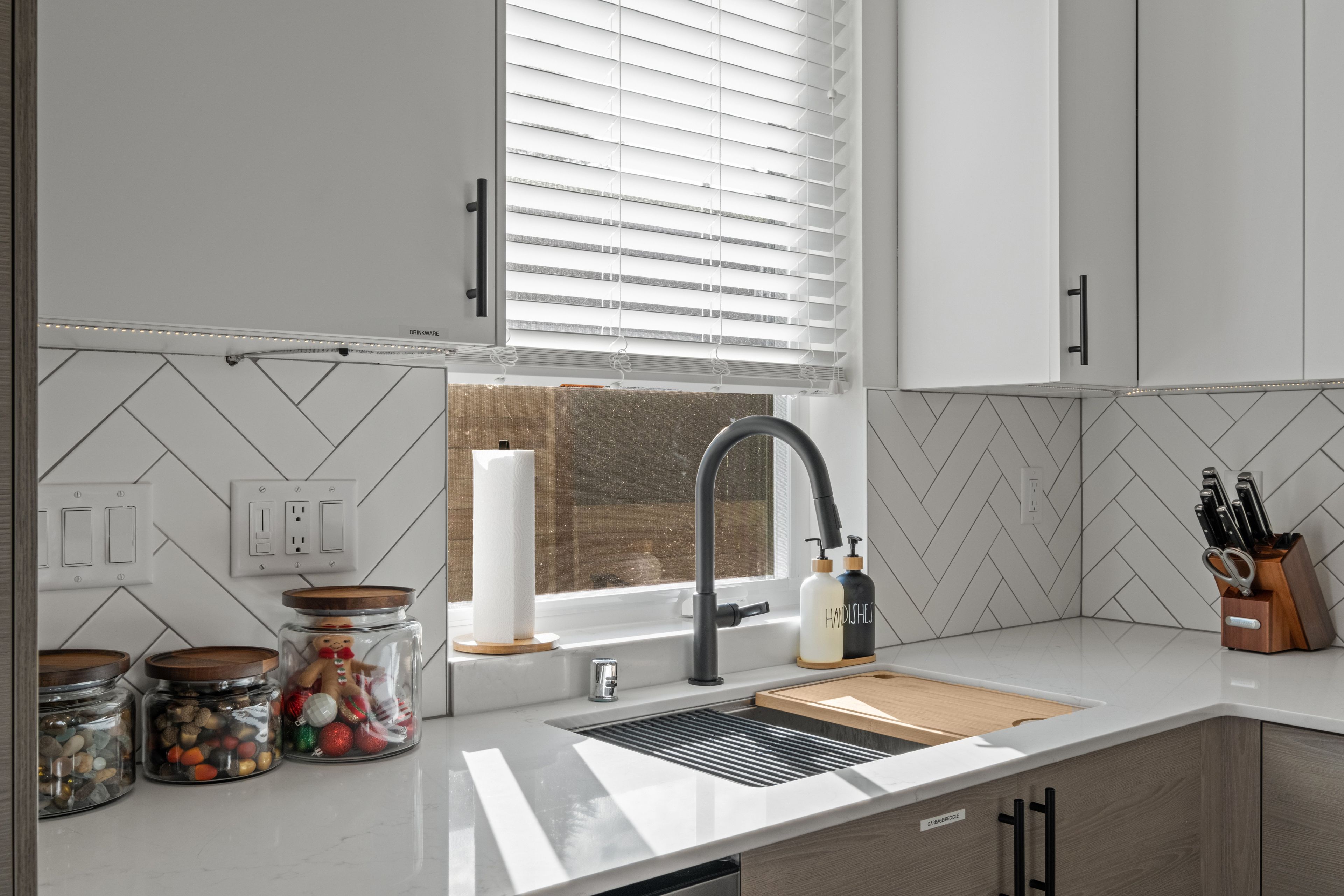 Une cuisine moderne équipée d’un évier, d’un robinet et d’un plan de travail. On y trouve des bocaux contenant des objets décoratifs, une fenêtre avec des stores et un bloc à couteaux.