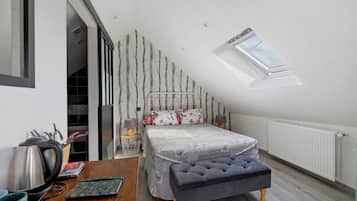 Ein Schlafzimmer mit schrägem Dach, einem Oberlicht, einem Bett mit floraler Bettwäsche, einem Nachttisch mit Wasserkocher und einer Heizung.
