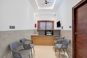 A reception area with a wooden reception desk, two grey chairs, a ceiling fan, a fire extinguisher, and a television on the wall.