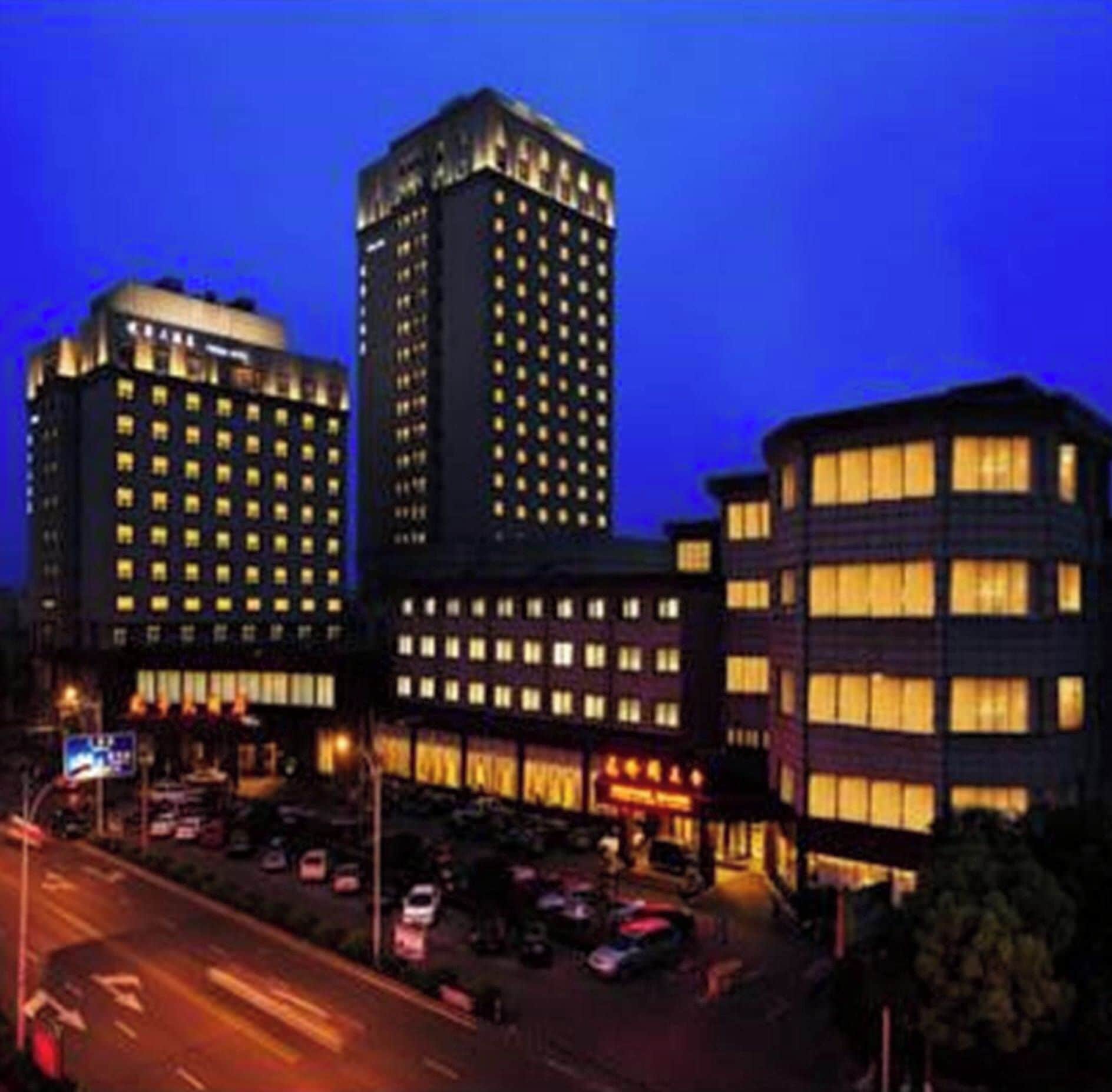 A multi-story building with illuminated windows, a parking lot, and a street with vehicles.