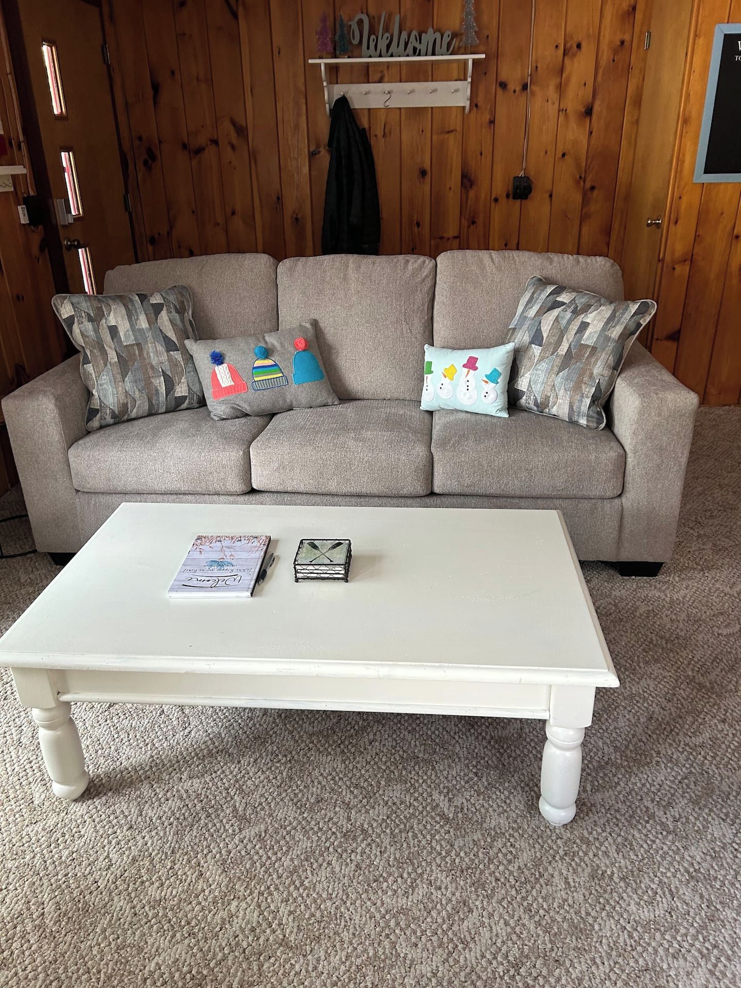 Un sofá color beige con cojines estampados, una mesa de centro blanca y un cartel que dice 'Welcome' en la pared.