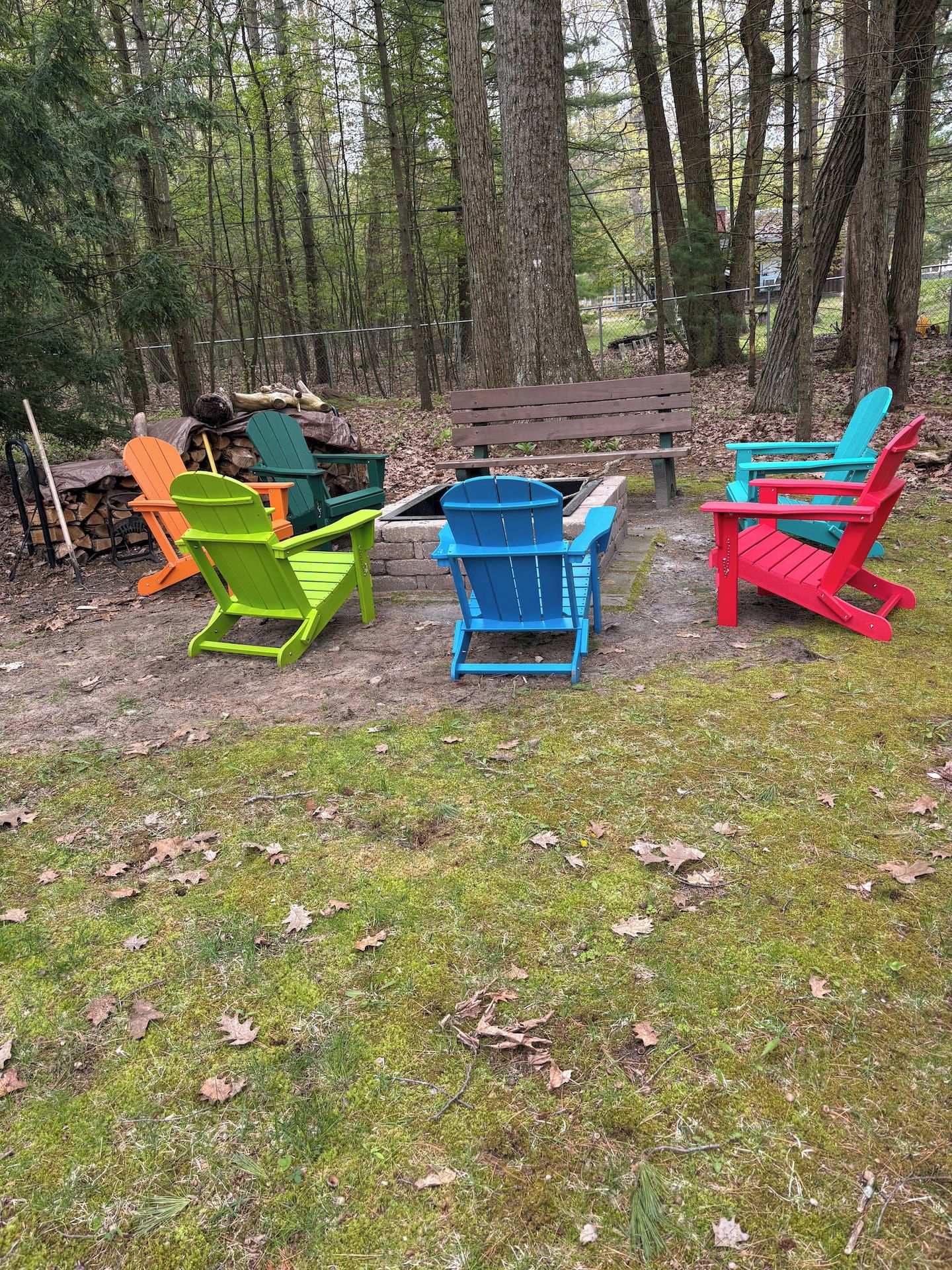 A forested area with a wooden bench and colorful Adirondack chairs.