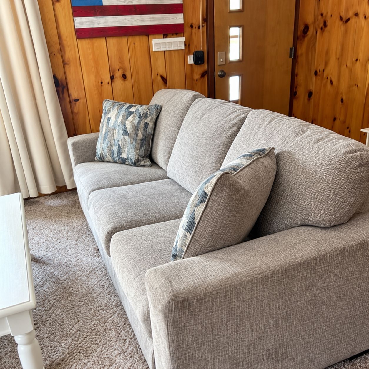 Ein graues Sofa mit gemusterten Kissen, ein weißer Tisch und eine Holzwand mit einer Flagge.