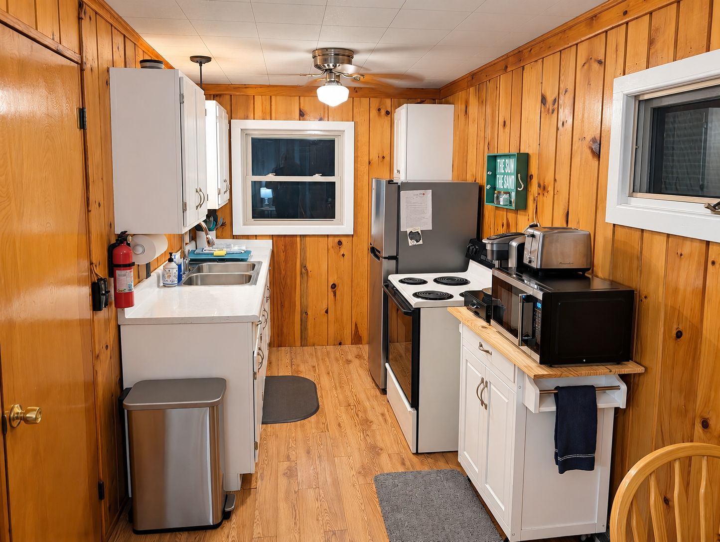 A compact kitchen with wooden walls, a window, a stove, a microwave, and a sink.
