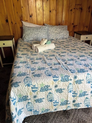A bed with a patterned bedspread, pillows, and a folded towel on a wooden floor.