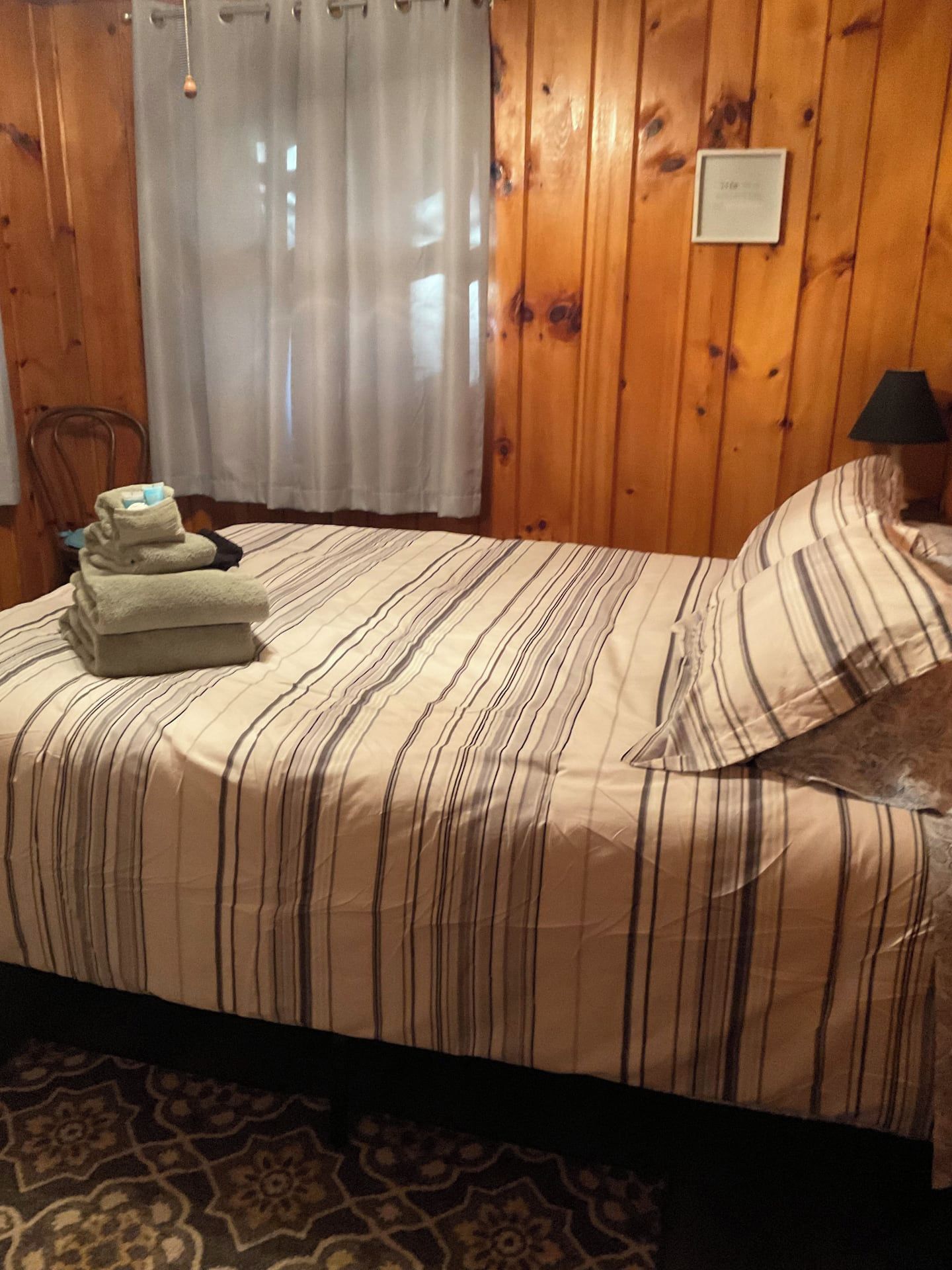 A bed with striped bedding, a folded towel stack, a lamp, and a wooden wall with a plaque.