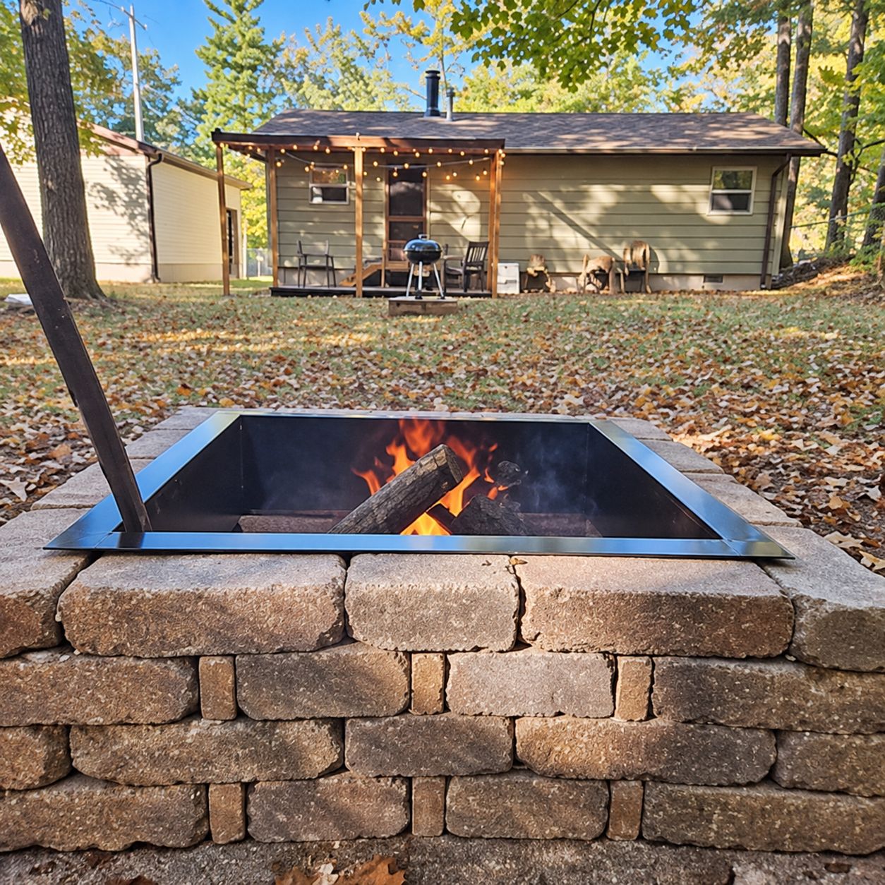 A fire pit with a brick surround, a house with a porch in the background, and a clear sky.