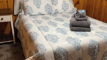 A neatly made bed with a floral patterned bedspread and pillows, a small bedside table, and a stack of folded towels.