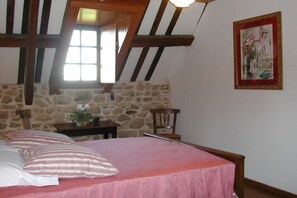 Ein Schlafzimmer mit Steinwand, Holzbalkendecke, einem Bett mit rosa Bettwäsche, einem Sessel, einem kleinen Tisch mit einer Vase und einem gerahmten Bild an der Wand.