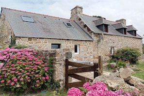 A stone building with a sloped roof, a wooden gate, and pink flowers in the foreground. - Holiday home in Crozon with swimming pool (crozon)