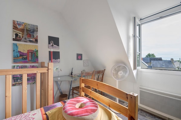 Una habitación con techo inclinado, una cama de madera con almohada, un escritorio con silla, un ventilador fijado a la pared y una ventana con vistas a edificios.