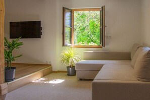 Ein Wohnzimmer mit einer Couch, einem an der Wand montierten Fernseher, einem Fenster mit Blick auf Grünflächen und zwei Blumentöpfen.