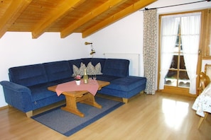 A room with a blue sofa, a wooden table, a radiator, a window with curtains, and a wooden ceiling. - Holiday apartment with 1 bedroom (Garmisch-Partenkirchen)