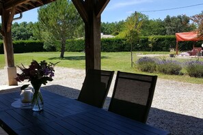 Uma área de estar ao ar livre com mesa e cadeiras, um vaso com flores e vista para um jardim com árvores e arbustos.