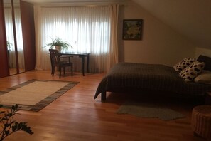 A bedroom with a bed, a desk, and a chair. There is a window with curtains, a plant, and a painting on the wall.