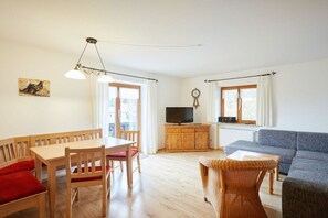Un salon comprenant un canapé, un téléviseur posé sur un meuble en bois, une table à manger avec des chaises et un fauteuil en osier.
