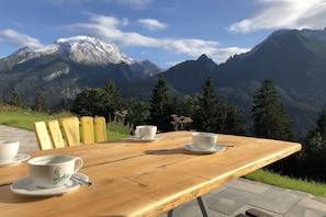 Ein Holztisch mit vier weißen Tassen und Untertassen, vor einer Berg- und Baumlandschaft.