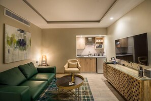 A modern living room with a green sofa, a coffee table, and a TV. There is a kitchen in the background with a microwave and a stove. - Ewaa Express Hotel - Gaber Riyadh (Riyadh)