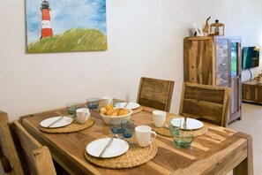 A dining table set with plates, cups, and a bowl of bread, with a wooden cabinet and a painting of a lighthouse in the background. - Seaside magic by the sea (Norddeich)