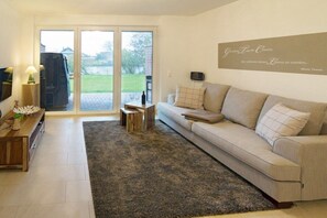 A living room with a sofa, a coffee table, a TV, and a sliding glass door leading to an outdoor area. - Seaside magic by the sea (Norddeich)