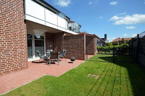 A residential building with a brick exterior, a covered patio with outdoor furniture, and a well-maintained lawn. - Holiday apartment with 2 bedrooms (Norddeich)