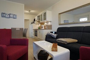 A living room with a sofa, a red armchair, a white coffee table, and a kitchen in the background. - Holiday apartment with 2 bedrooms (Norddeich)