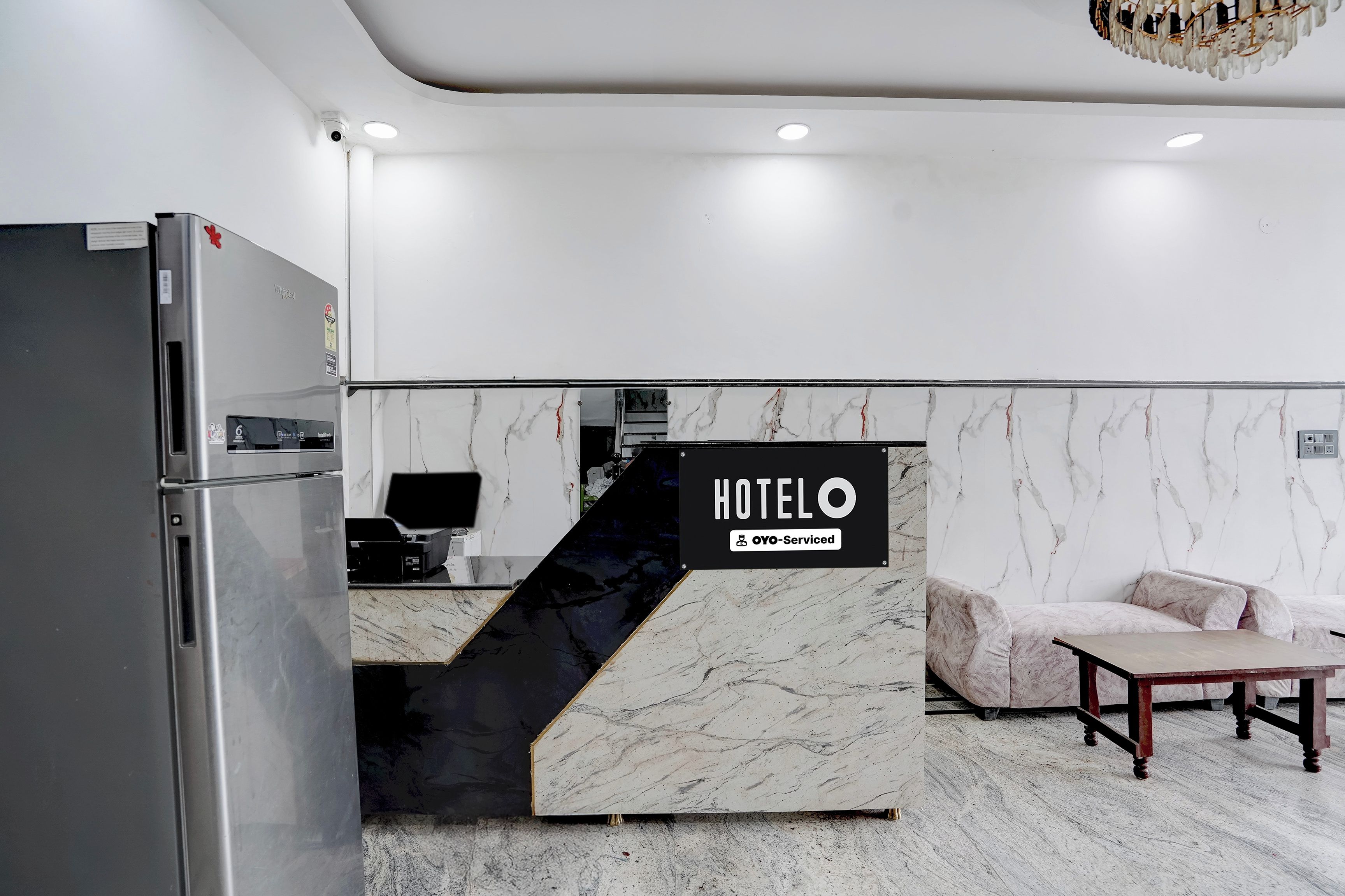 Zona de recepción de hotel con pared de mármol, mostrador de recepción, refrigerador, sofá y una pequeña mesa.
