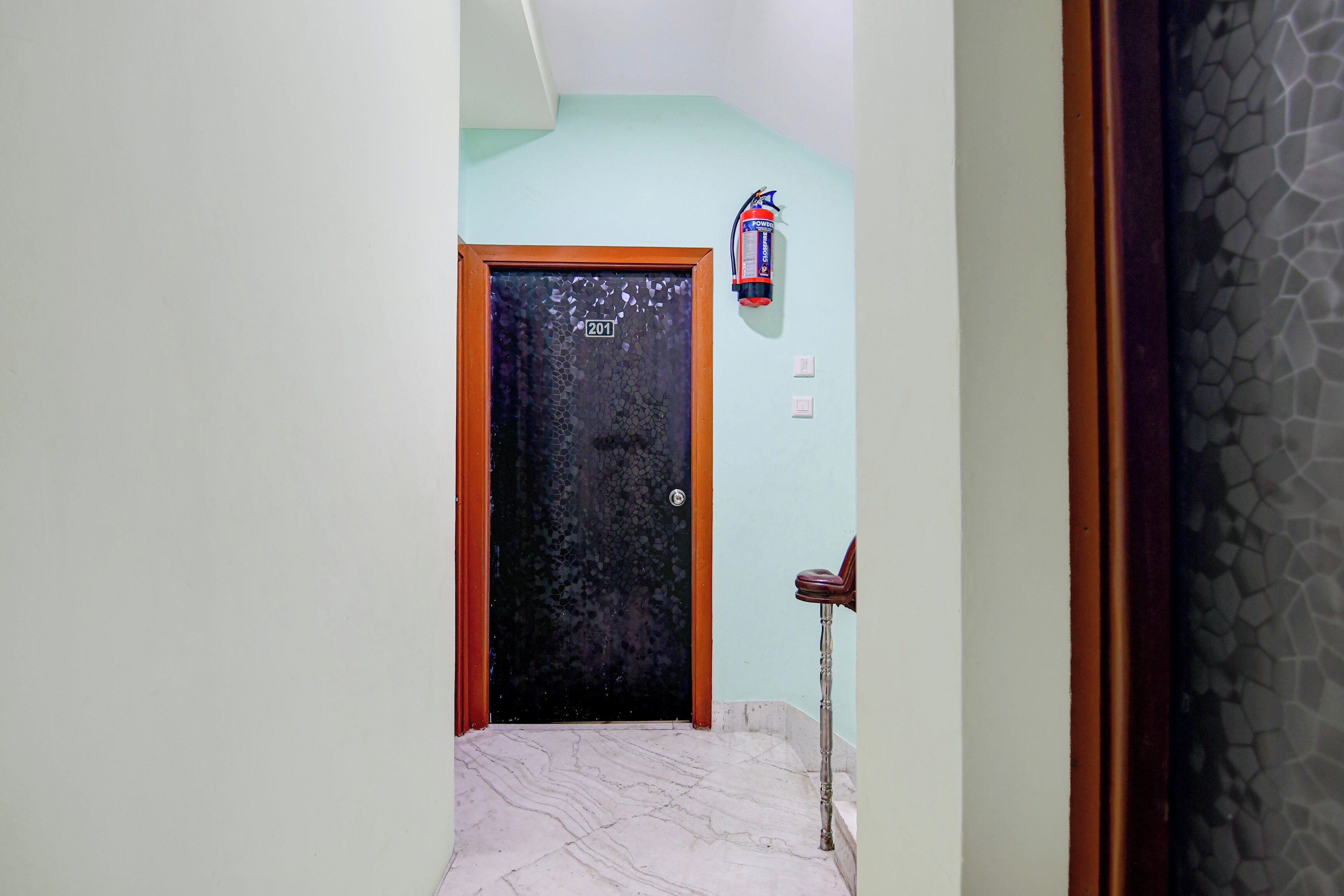 A hallway with a tiled floor, a door with a number sign, a fire extinguisher, and a wall with a textured pattern.