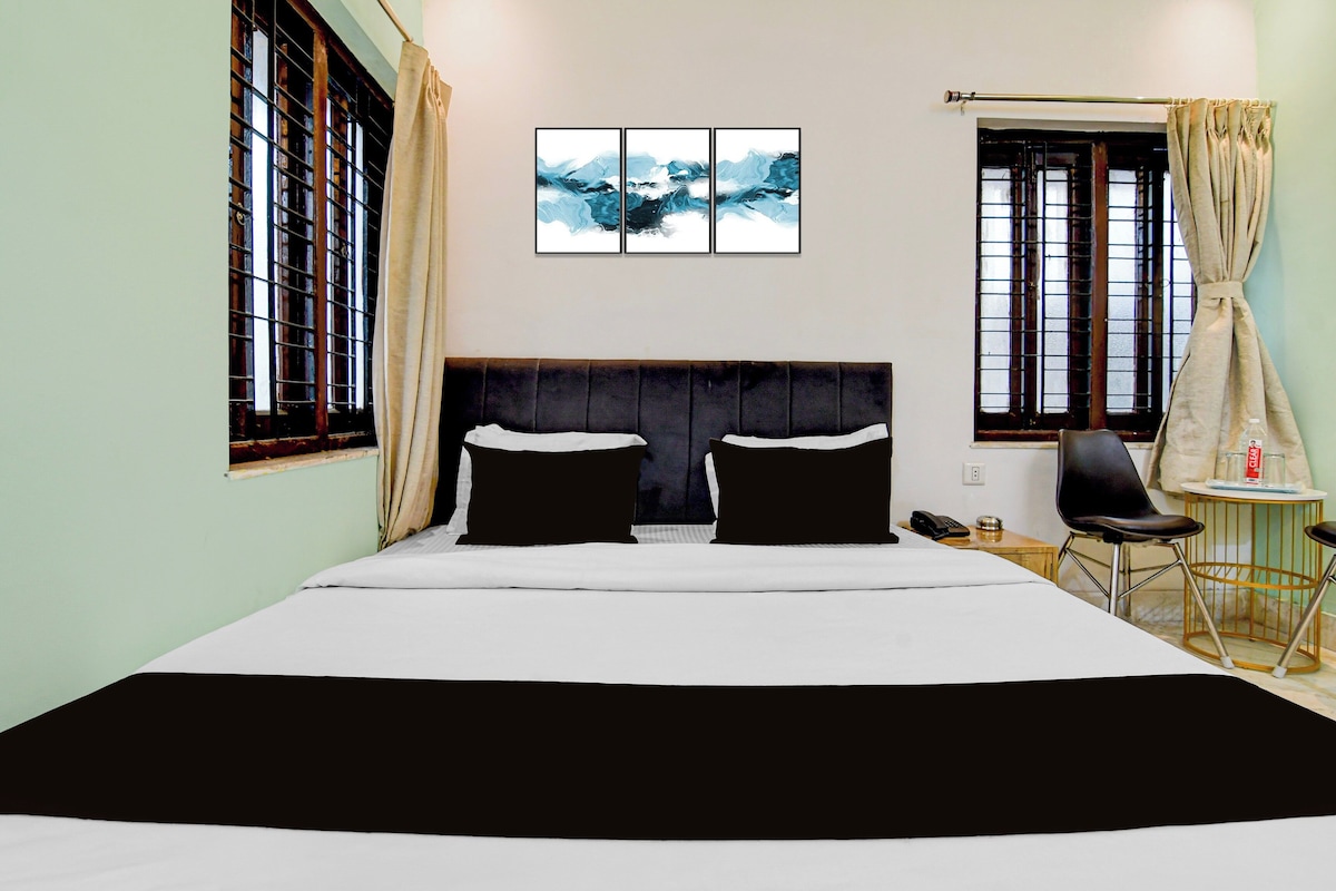 Quarto de hotel com uma cama grande, duas almofadas pretas, cabeceira, uma mesinha redonda com cadeira, telefone e um quadro na parede.