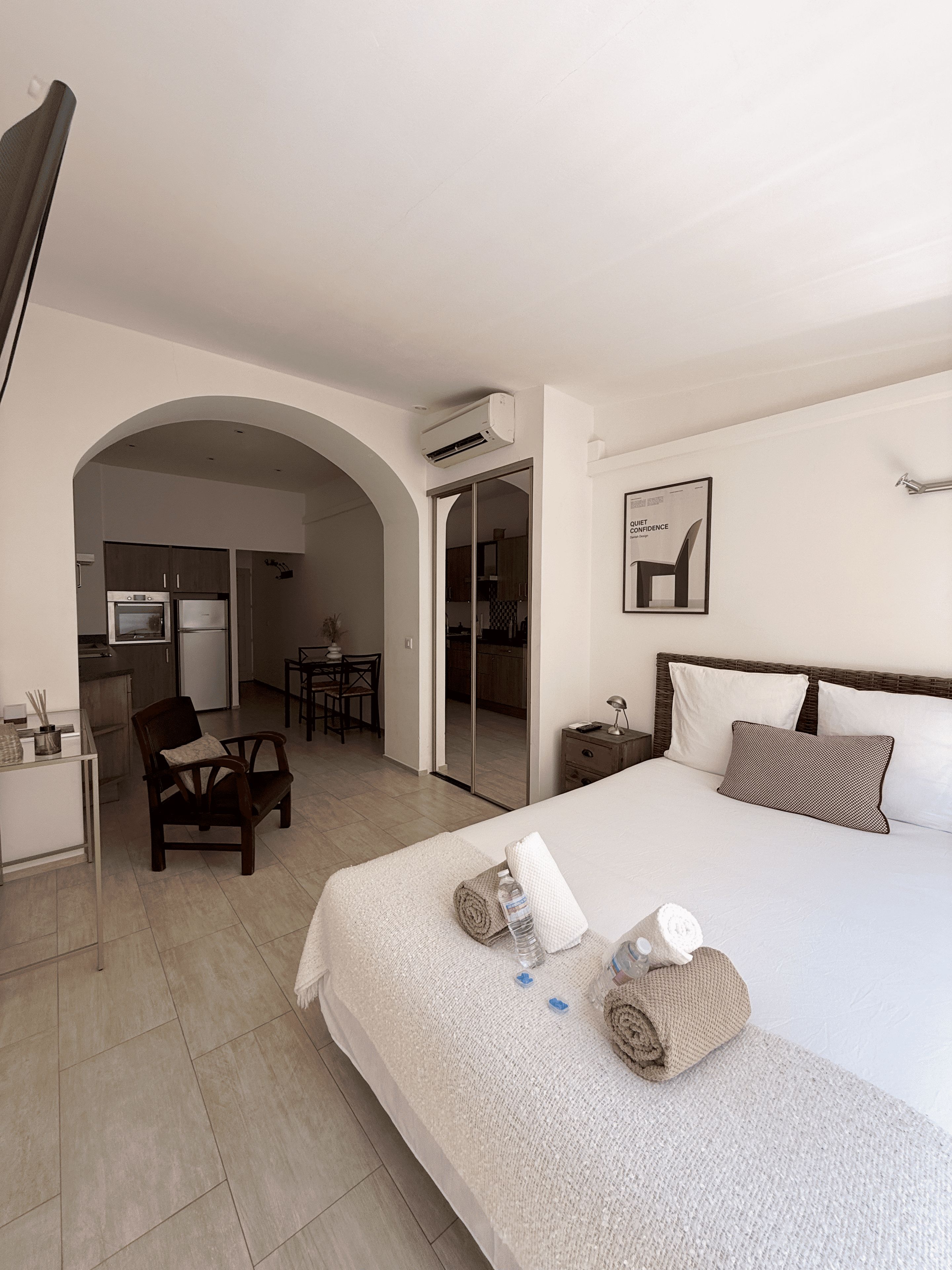 Une chambre d’hôtel comprenant un lit, un bureau, une chaise et une porte en arc donnant sur un coin cuisine.