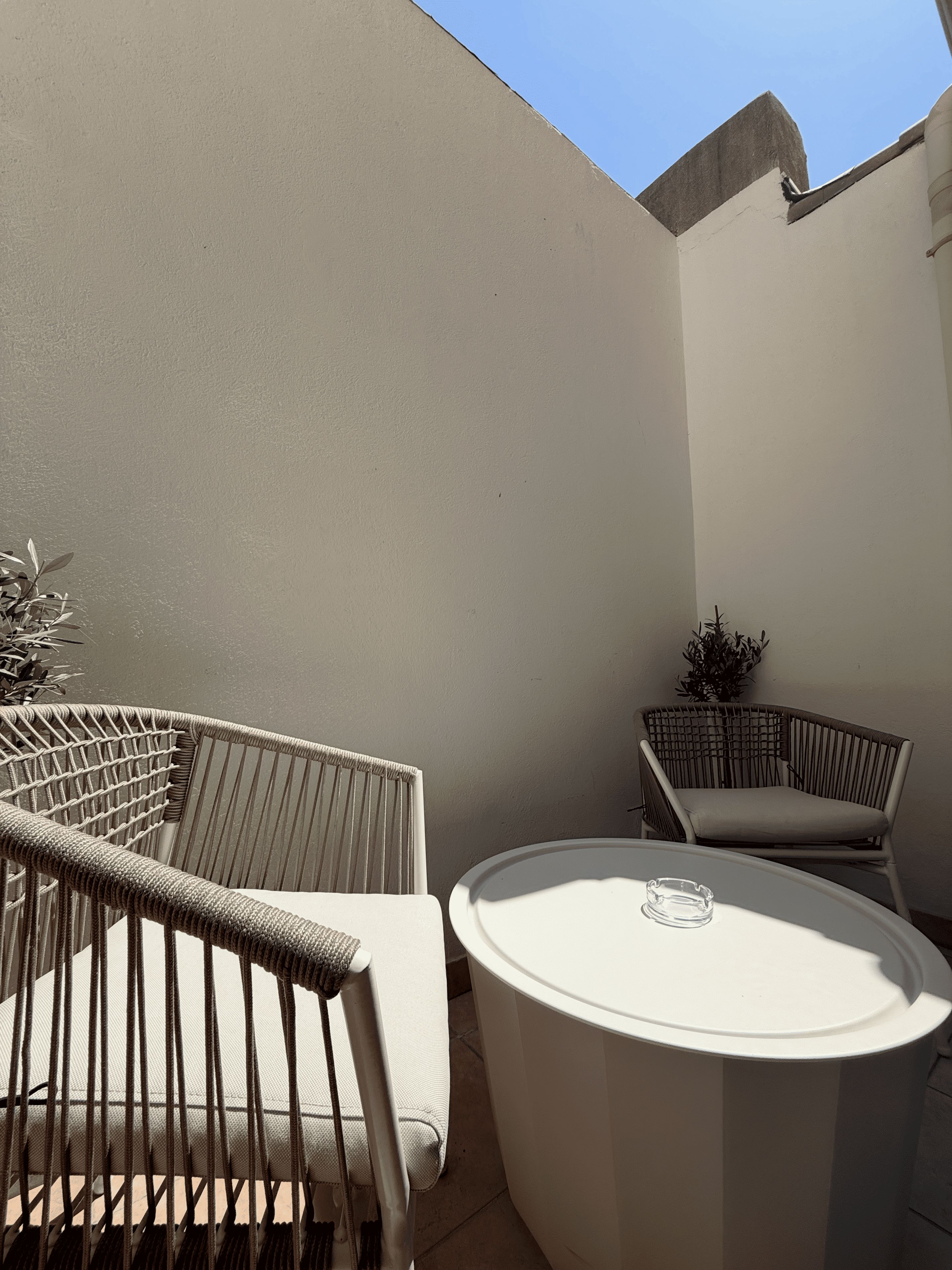 Un petit espace extérieur aménagé avec deux chaises en osier et une table ronde, entouré d’un mur sobre et d’un ciel dégagé.