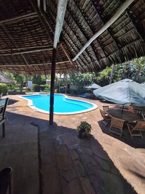Un espace piscine doté d’un toit de chaume, d’un sol en pierre et de mobilier en bois.