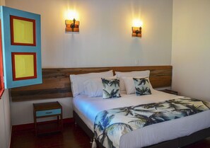 Un dormitorio con cabecera de madera, cama con ropa de cama blanca y almohadas estampadas con motivos de hojas de palma, dos luces fijadas a la pared y una mesita de noche con cajón.