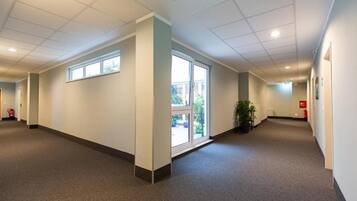 A hallway with a carpeted floor, white walls, and ceiling lights. There are windows on the left side and a door at the end.
