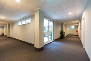 Un pasillo con piso alfombrado, paredes blancas y luces en el techo. Hay ventanas a la izquierda y una puerta al final.