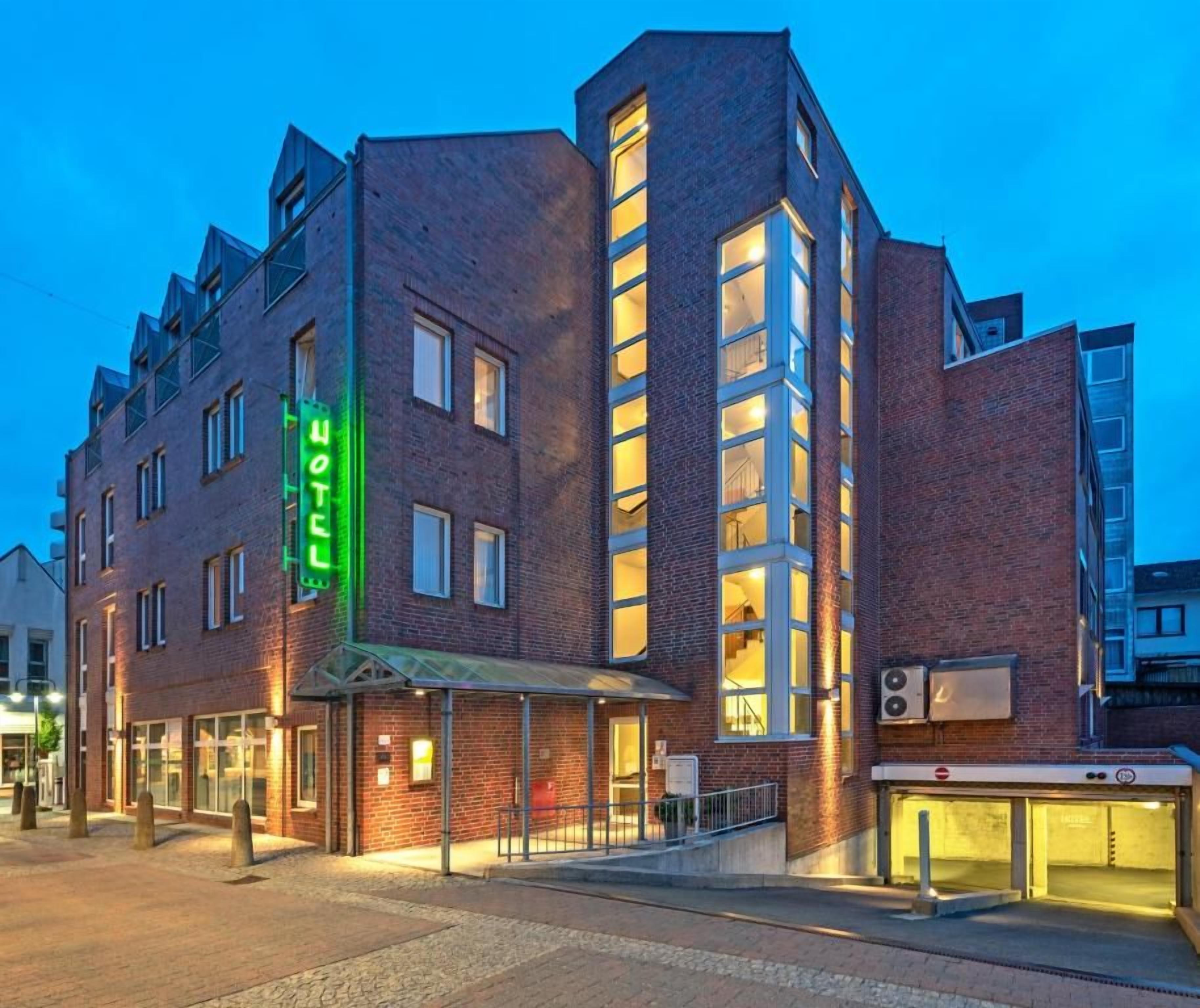 Un hôtel moderne, reconnaissable à son grand panneau vert, à son entrée abritée et à son ascenseur bien visible.