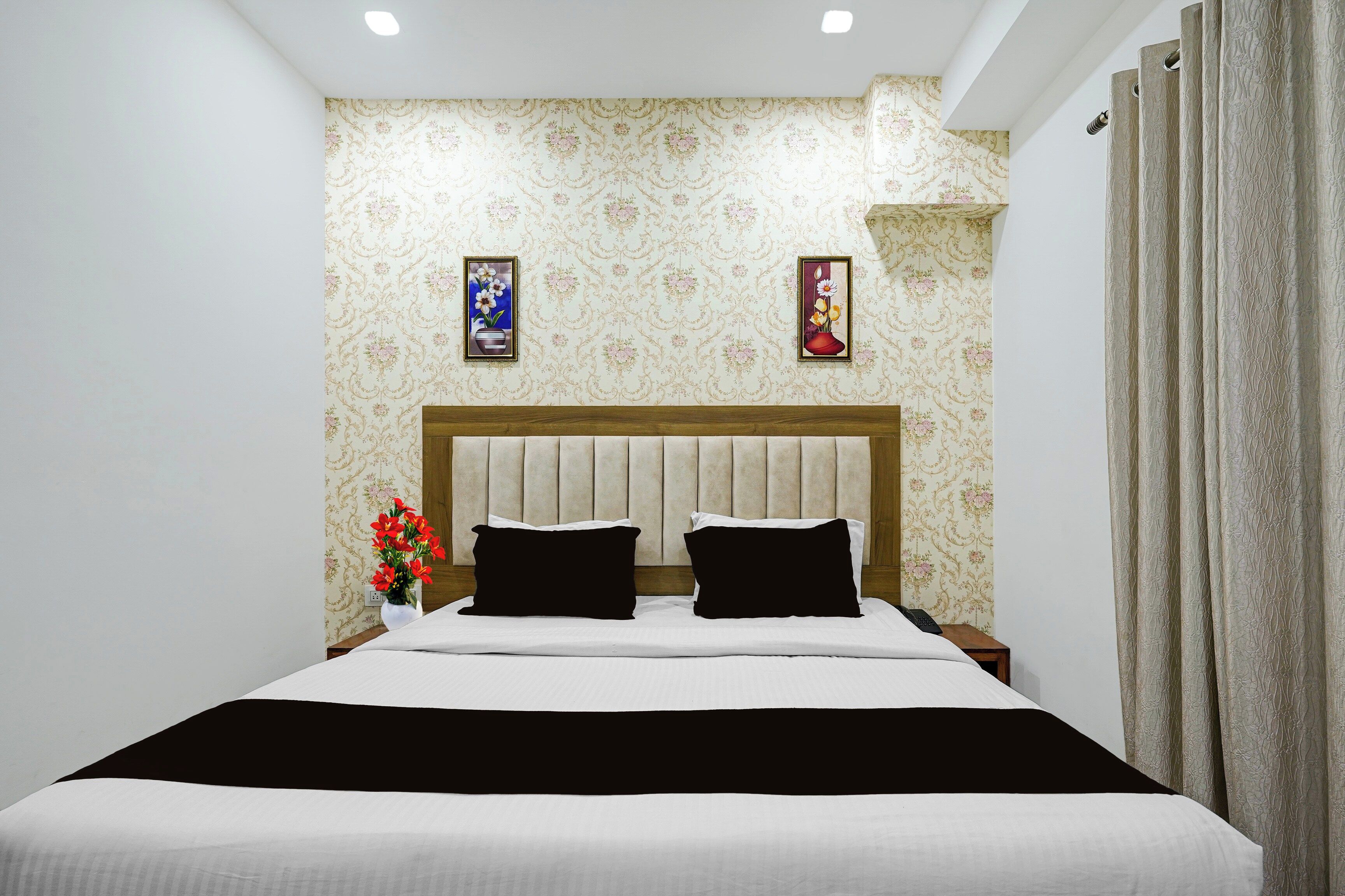 Un dormitorio con una cama, dos almohadas, una mesita de noche, un florero con flores y dos cuadros colgados en la pared.