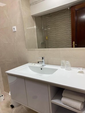 Un baño con un lavabo blanco, un espejo encima y una ducha visible en el reflejo.