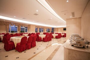 Un salón de banquetes con mesas redondas y sillas rojas, una zona de buffet y grandes ventanales.