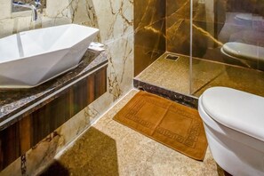 A bathroom with a white vessel sink, a marble vanity, a glass shower enclosure, and a toilet with a brown mat.