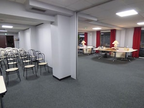Sala de conferências com mesas, cadeiras, divisória de vidro e cortinas vermelhas.