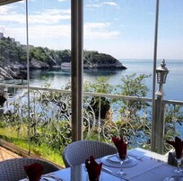 A dining area with a table set for two, overlooking a body of water and a rocky coastline.
