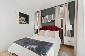 A modern bedroom with a large bed, a red headboard, two bedside tables with lamps, and a wall-mounted artwork.